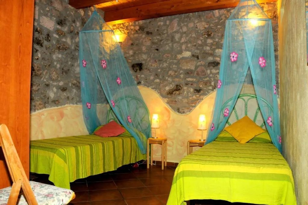 Monolocale di Su'e Lentu con due letti, copriletti verdi a righe, zanzariere azzurre a fiori e parete in pietra a vista