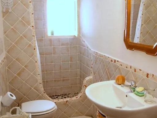 Bagno di Su'e Lentu, piastrelle beige a rombi, doccia in muratura e ciottoli sotto una finestra, lavabo bianco