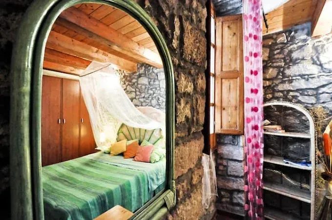 Specchio ad arco verde che riflette un letto con zanzariera e cuscini colorati in una camera di Su'e Lentu con muri in pietra